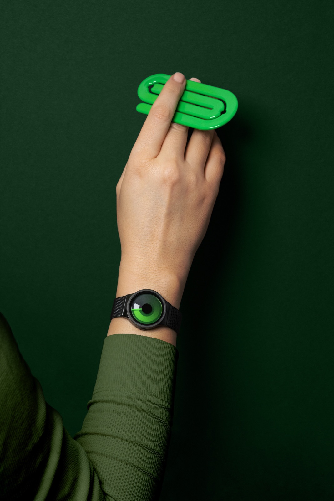 Hand with an oversized green paperclip and a ZIIIRO watch on dark green